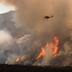 A helicopter flies closely over a wildfire on a mountain. You can see orange flames amongst a large cloud of white smoke. 