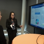 A researcher stands and points to screen showing her research poster. She is talking about the research with two people looking at the screen.