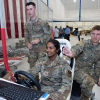 Three cadets are photographed; one is operating a steering wheel, controlling a robot off screen. The other two look on.