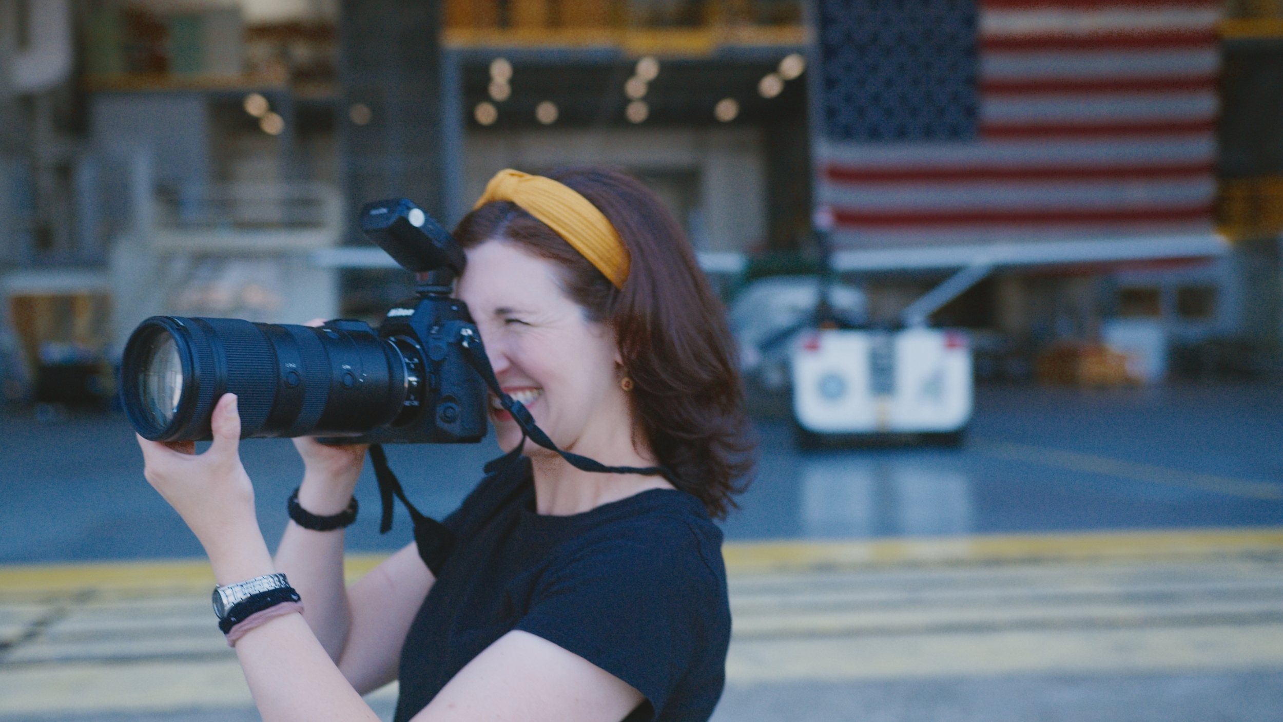 A woman smiles as she takes a photo with a professional camera, with a large American flag in the background.