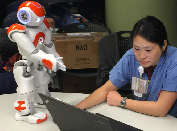 A nurse sits at a table, on top of which there is a laptop and an orange and white robot about 2 feet in height. 