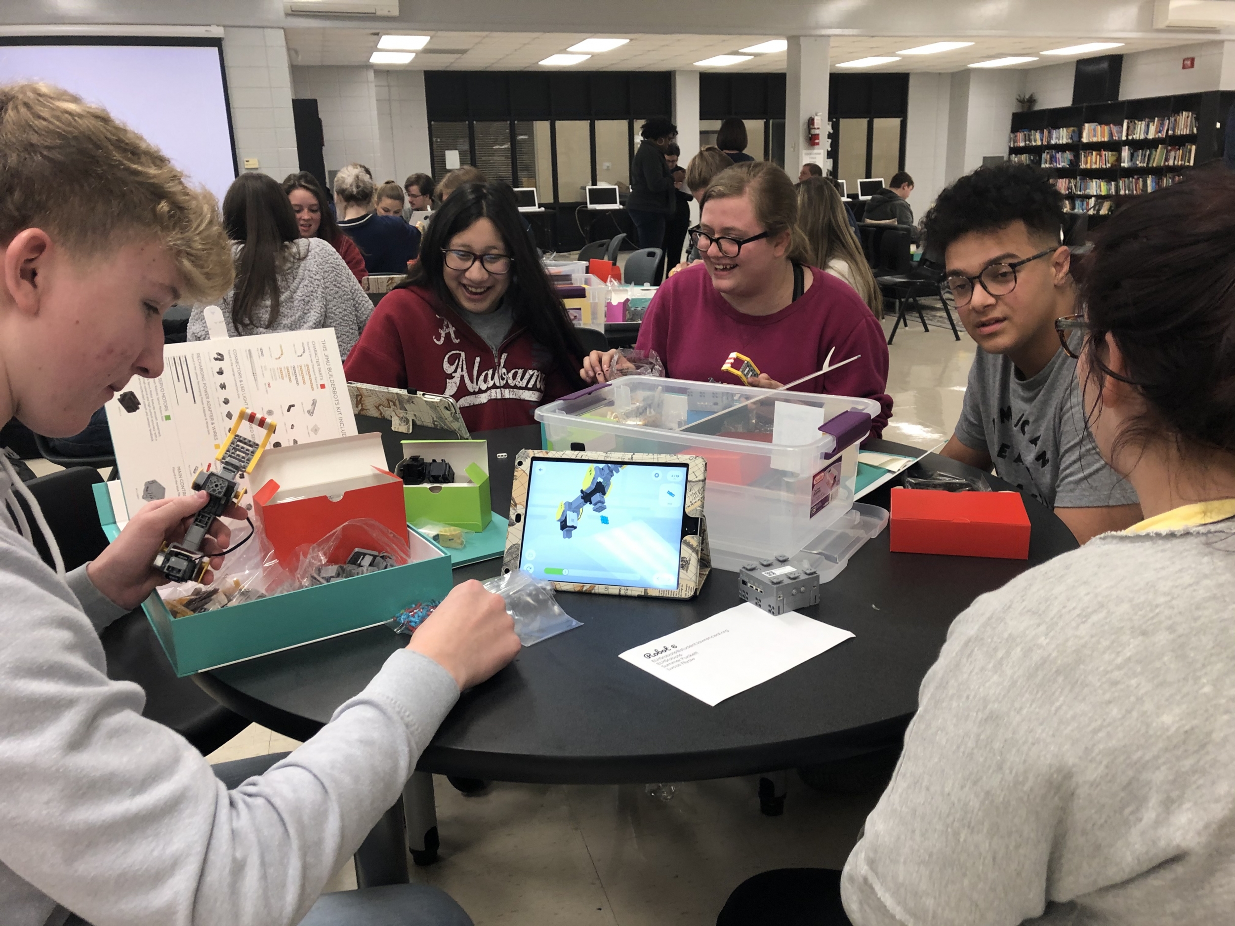A group of five high school students sit at a table, smiling, working together to assemble a robot out of what look to be lego-like parts. 