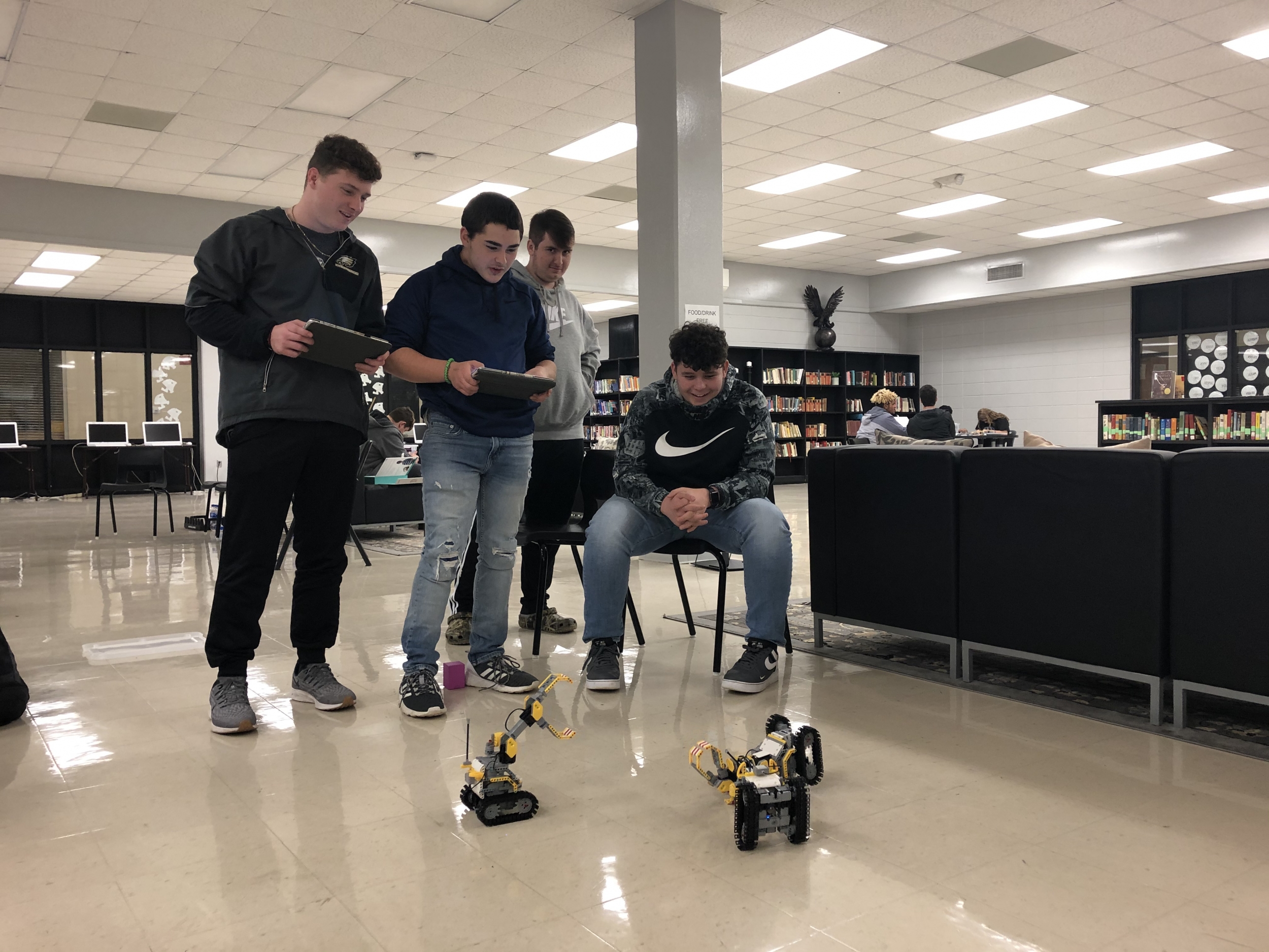 Four high school students look at 2 small robots on the ground that are moving, while one controls it from an ipad