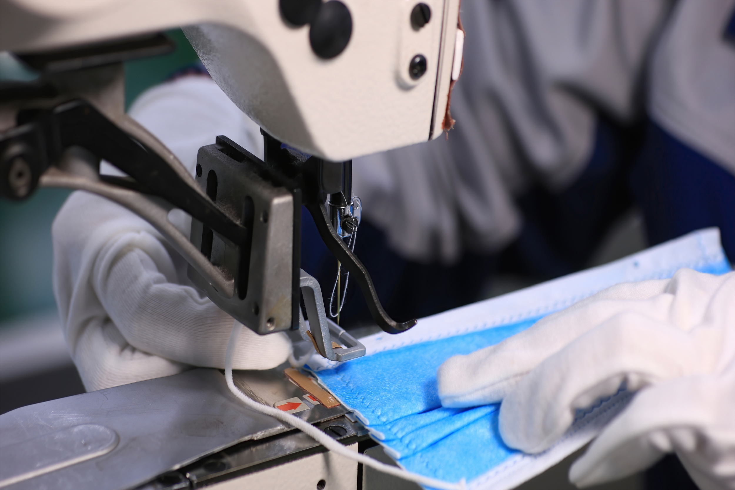 An image of a person (only their hands are shown) using a sewing machine to sew a blue surgical mask.