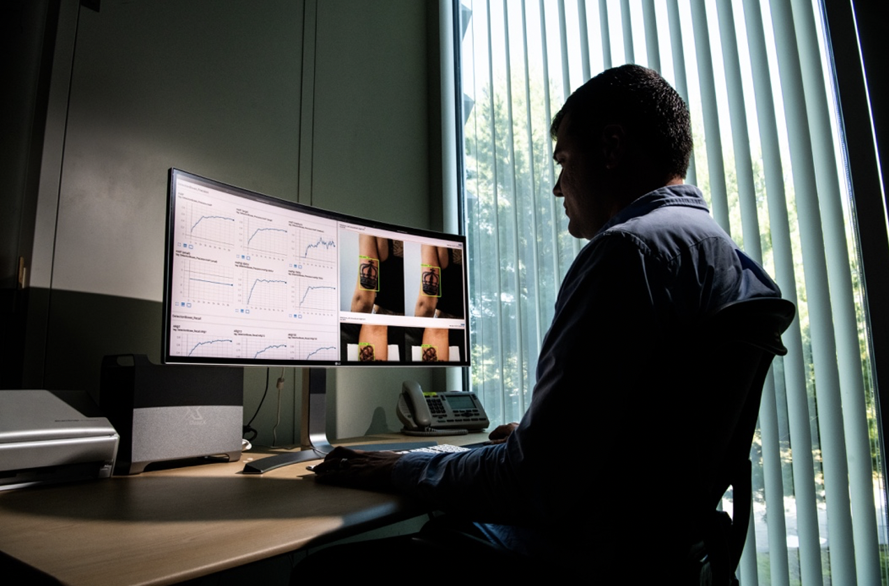 A man sits at an office desk looking at a large monitor. the left hand side shows data graphs, the right side shows two images of the back of a woman's arm with a crown tattoo, and a green box around the tattoo. 