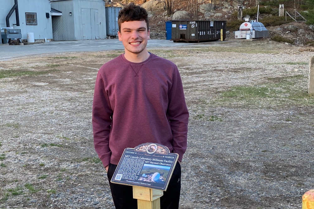 Scout Carter Purple stands outside in front of the plaque he made containing information about the Millstone Hill Radar and Haystack Observatory