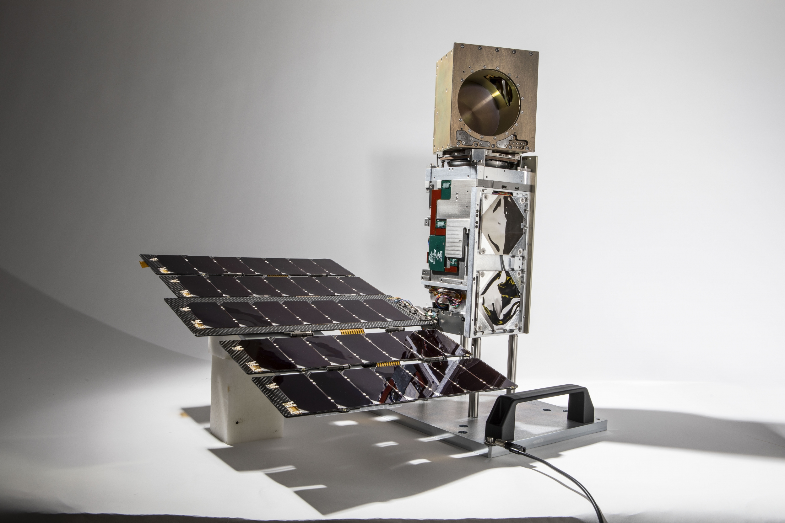 A small satelitte photographed against a white background. About , about 10 cm X 10 cm X 36 cm in size, it consists of a small solar panel array, connected to the satellite bus standing upright, topped with a golden cube.