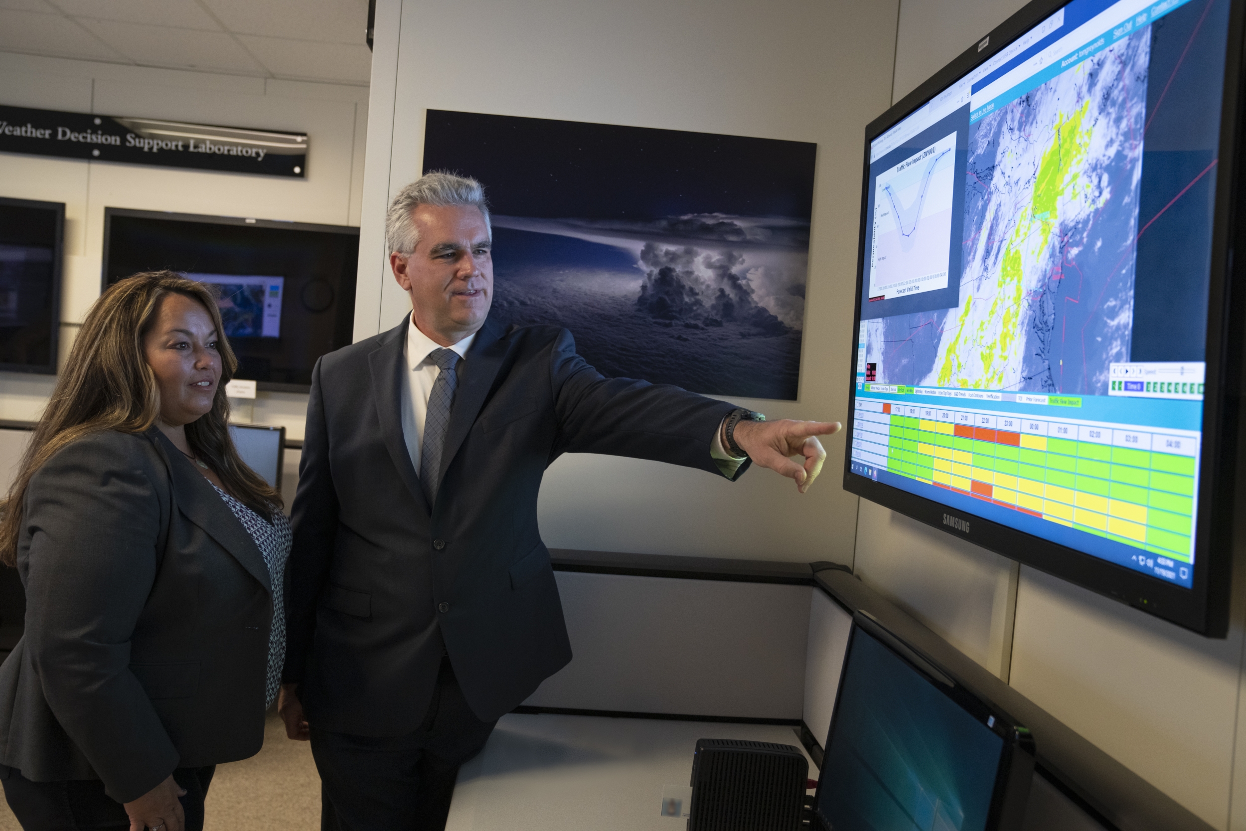 A man and woman stand next to a large monitor, looking at the screen, which is displaying a radr map and a green/yellow/red matrix. 