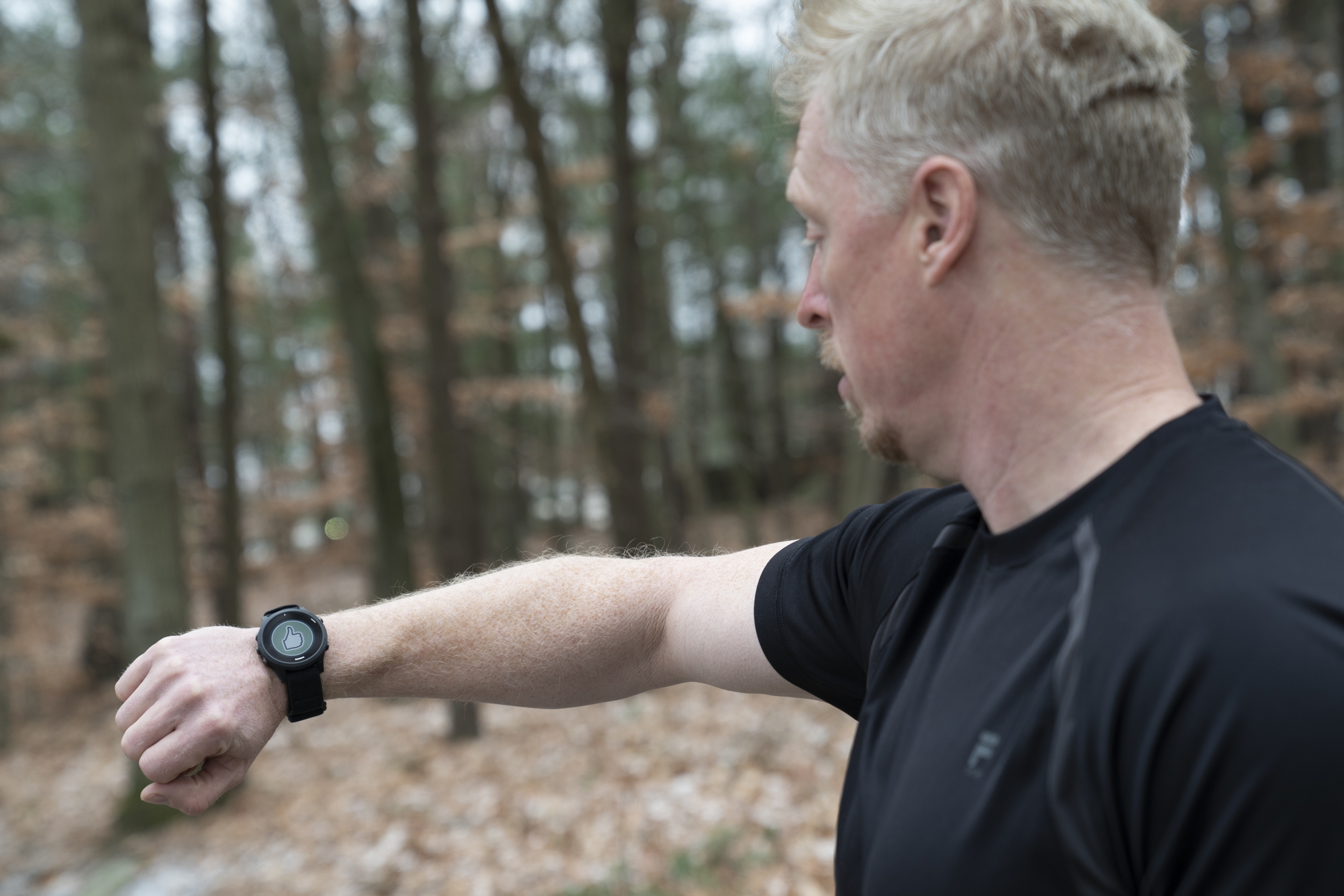 A man in black t-shirt looks at a smartwatch on his wrist, which is displaying a green circle with a thumbs up sign. The man is outside, with trees and leaves in the background.