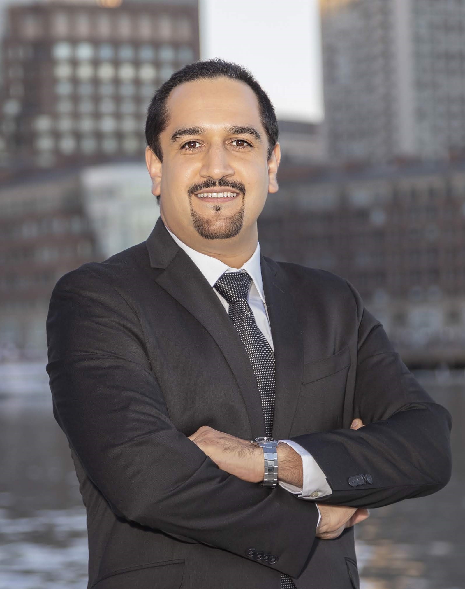 A photo of Hamed Okhravi. He is wearing a suit, with his arms crossed, with city buildings in the background. 