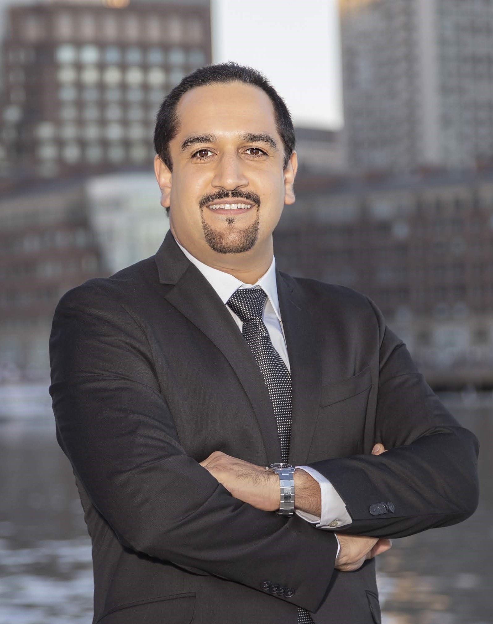A photo of Hamed Okhravi. He is wearing a suit, with his arms crossed, with city buildings in the background.