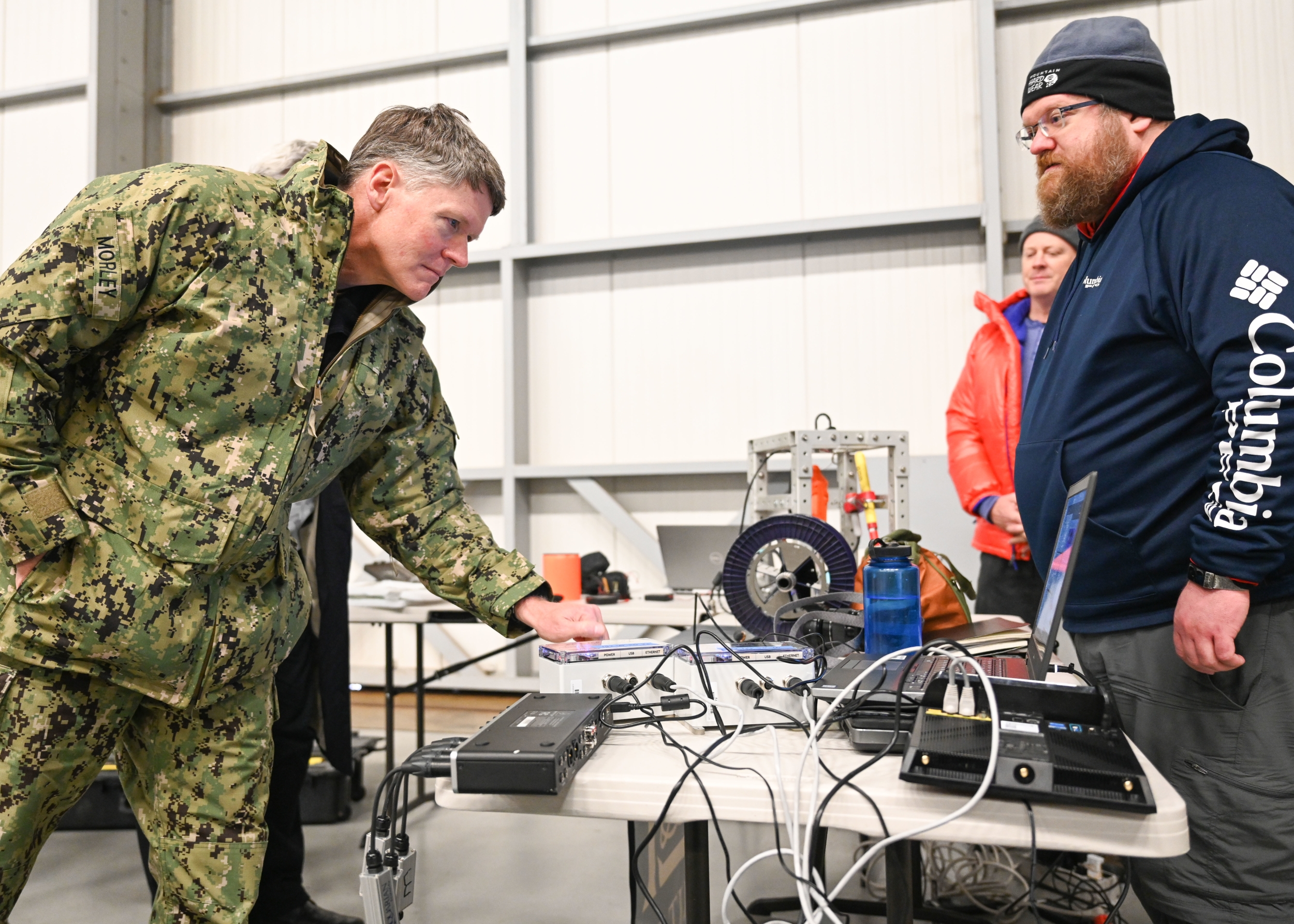 A military official looks at sensors on a table, across from a Lincoln Laboratory researcher. They are in large room, similar to a warehouse setting.  