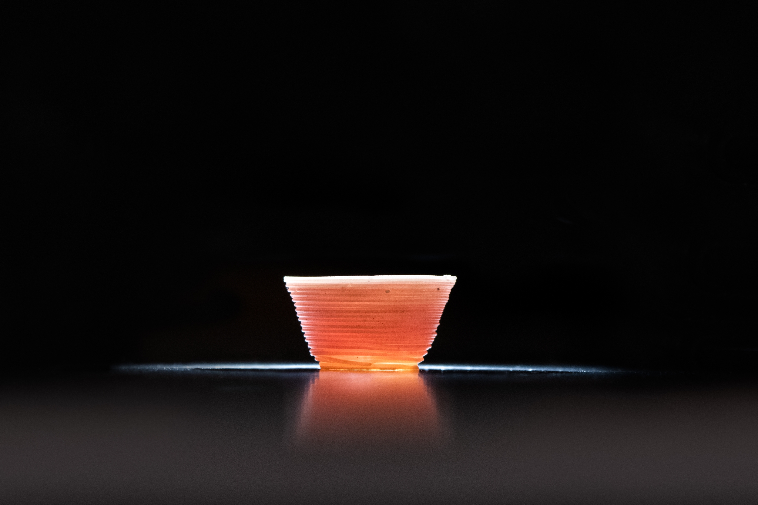 A small cup, made out of 3D printed glass, is lit from within, making the cup glow orange.