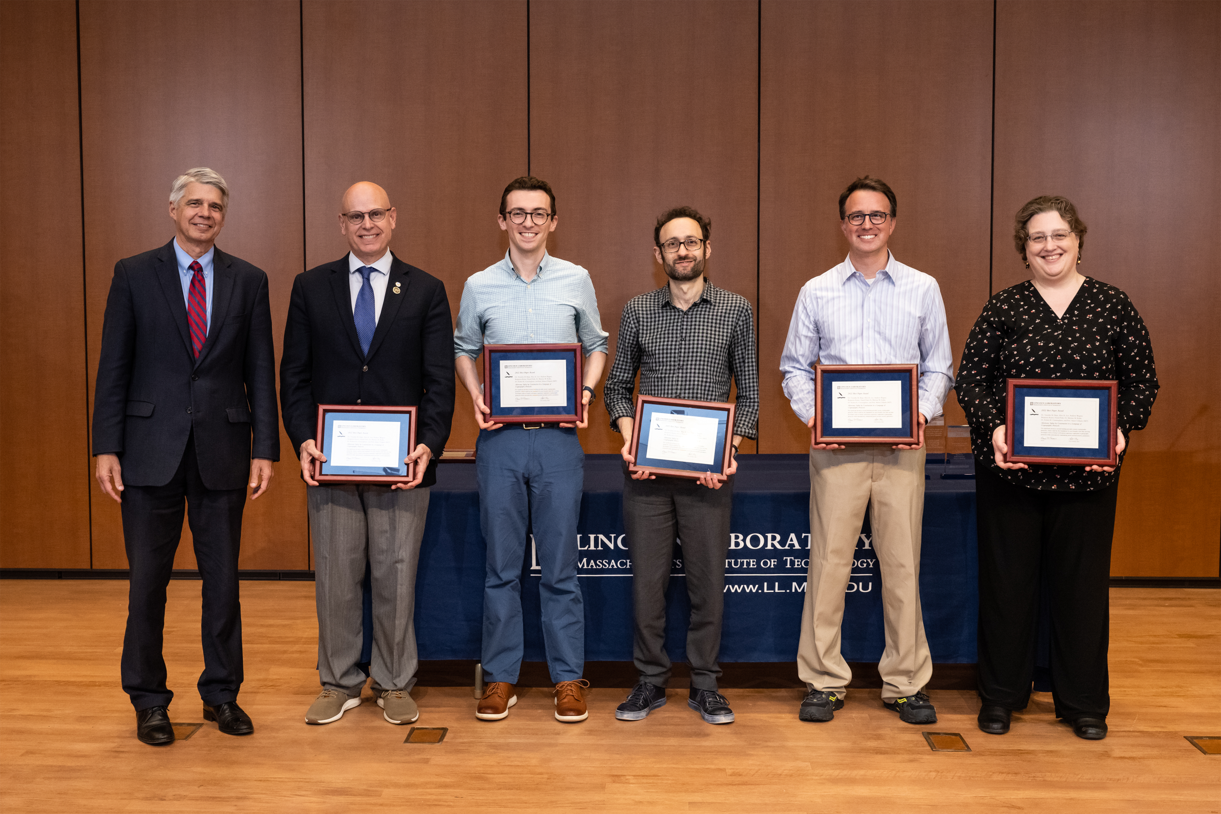 A group of six people pose a group photo on a stage. Five of the people hold award plaques.