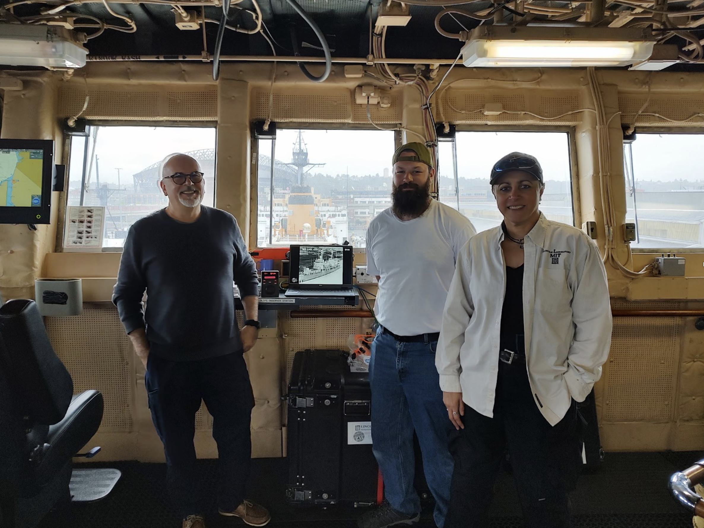 A group of three researchers standing inside the bridge of the healy boat. a computer screen is visible behind them showing imagery from their system.