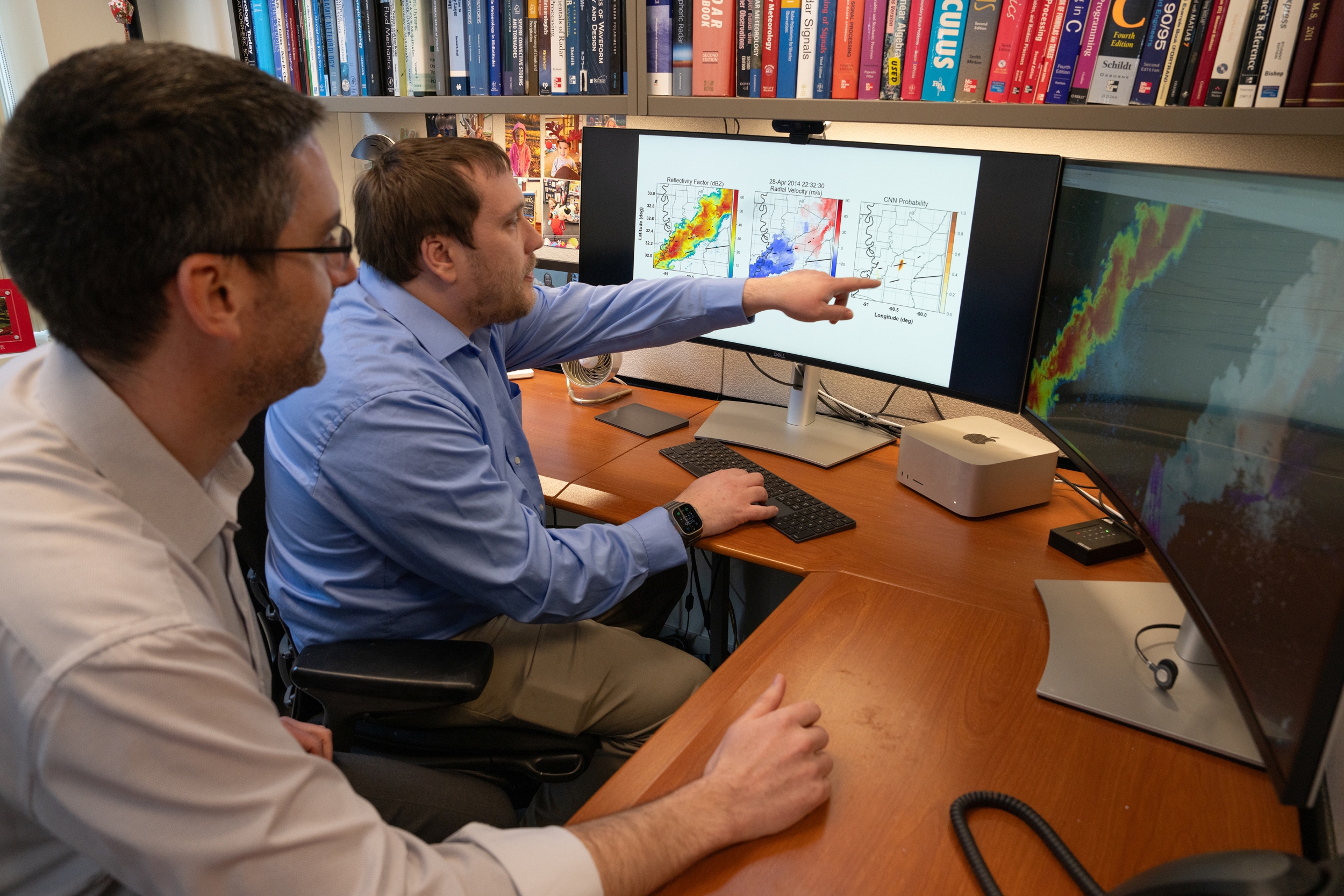 two scientists sit at a desk in front of two computer screens; the screens show radar images of tornadoes.