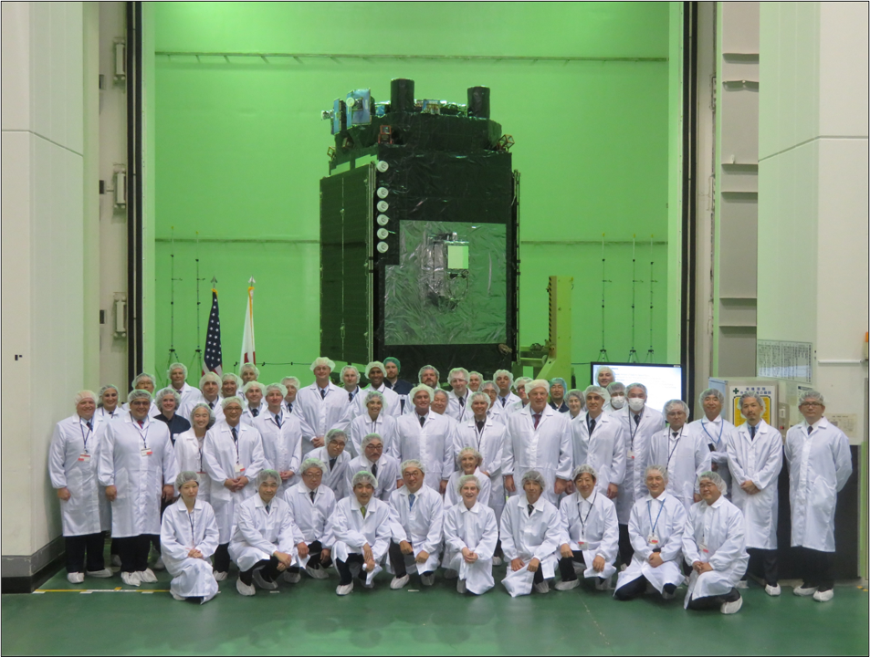 The group is wearing white lab coats and hairnets and are posed in a greenish room with the payload in the background.