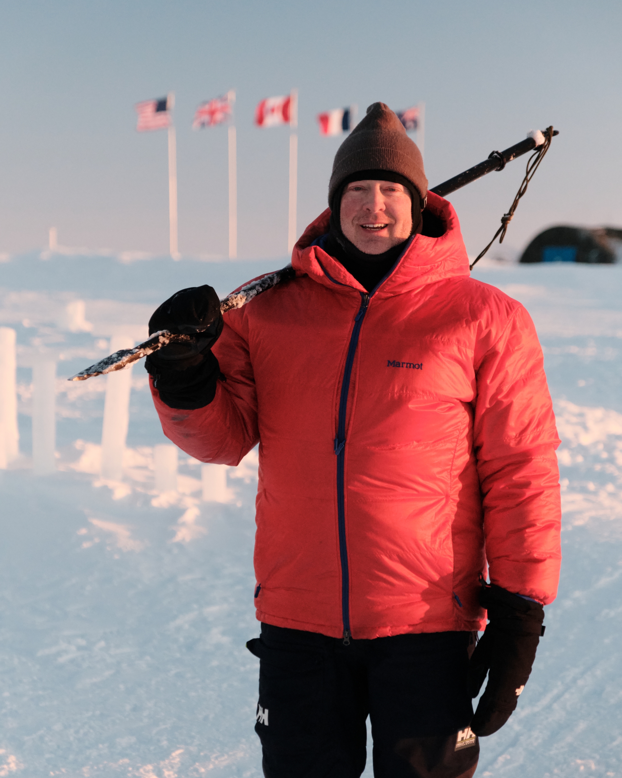 A man stands on ice in the Arctic with a walking stick flung over his shoulder; flags representing different nations are pictured in the background.