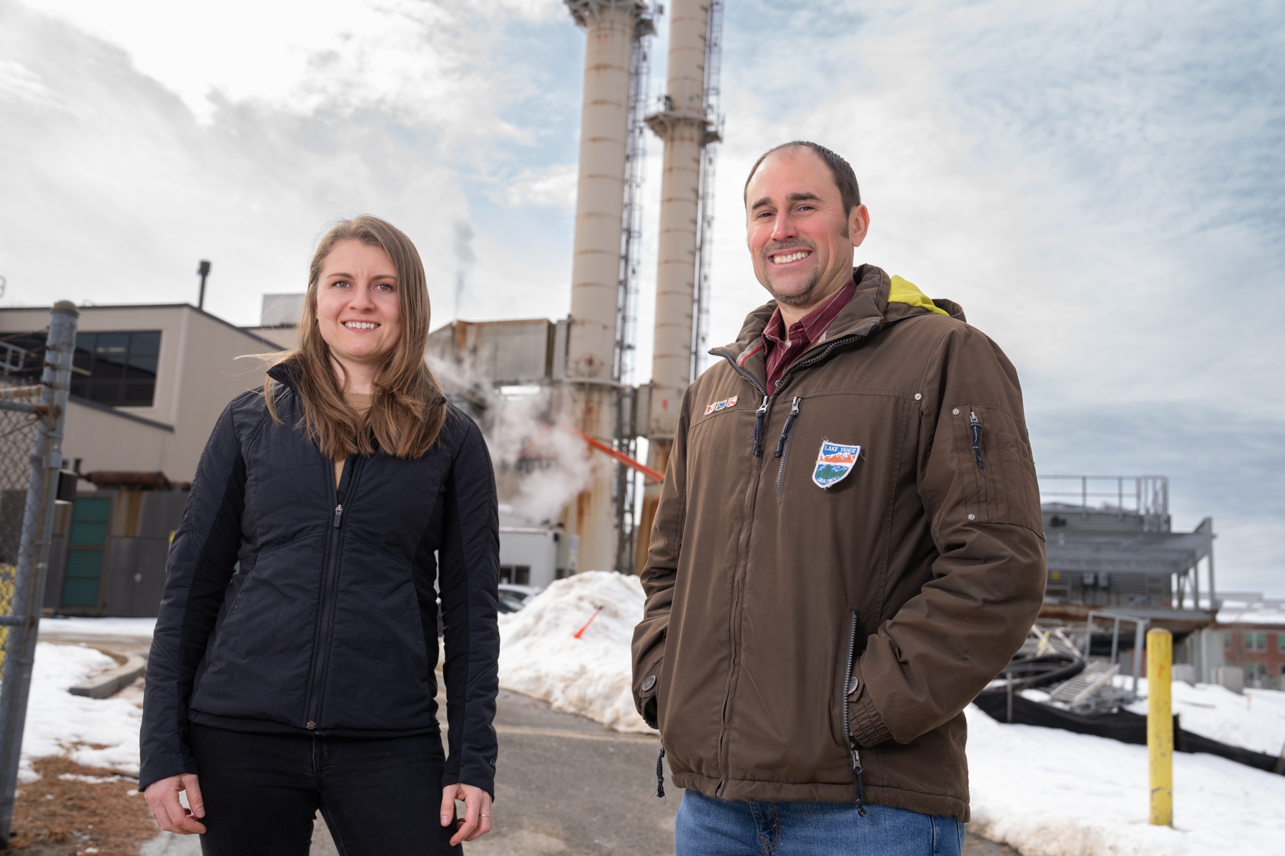 Jean Sack and Chris Lashway pose for a photo outside, with power-plant infrastructure in the background.