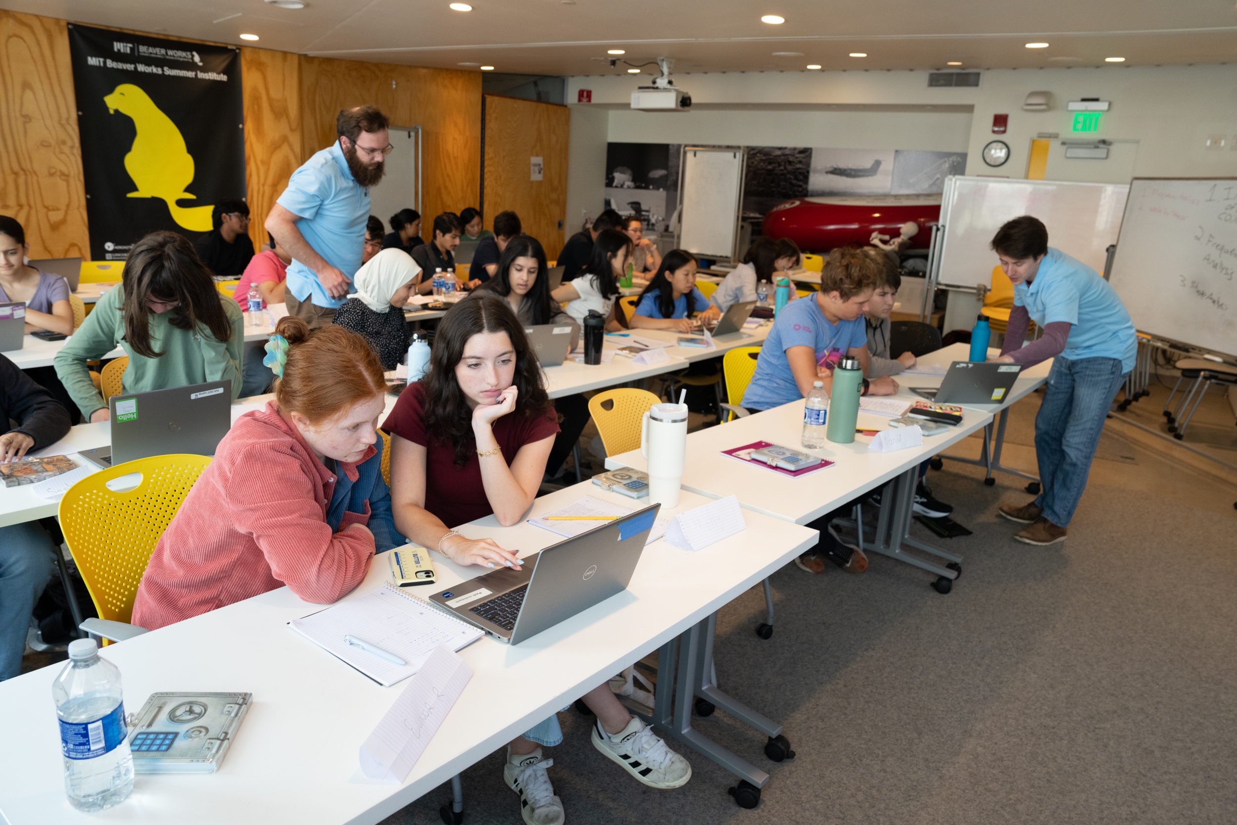 LLCipher students sit in groups of two at long tables working on laptops. Two instructors are helping the students.