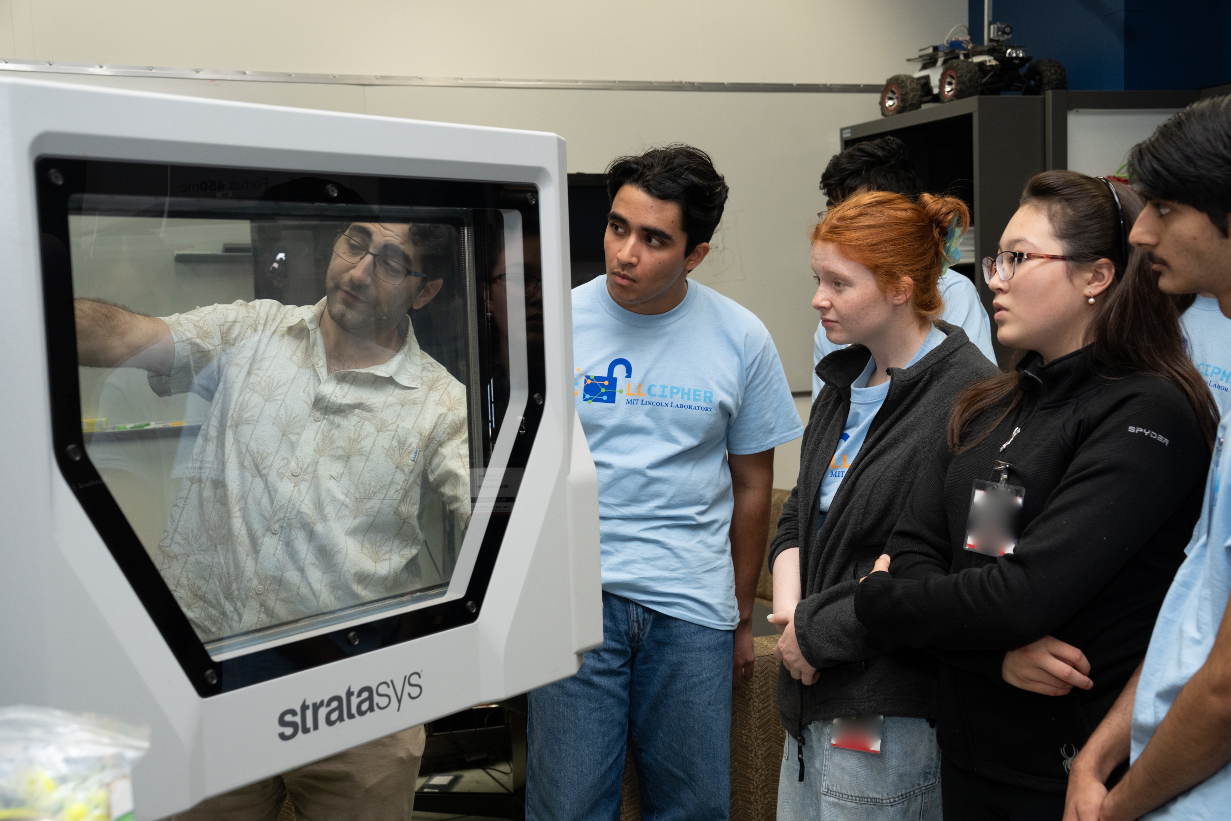 LLCipher students gather to learn about the Laboratory's 3D printer. A Laboratory staff member has the door of the printer open and is pointing inside while explaining.