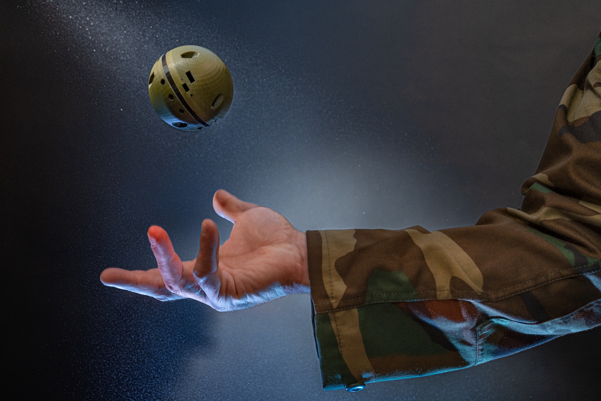 The TOSSIT sensor, a baseball-sized 3D printed sphere, is tossed in the air by a person offscreen wearing a camo jacket.