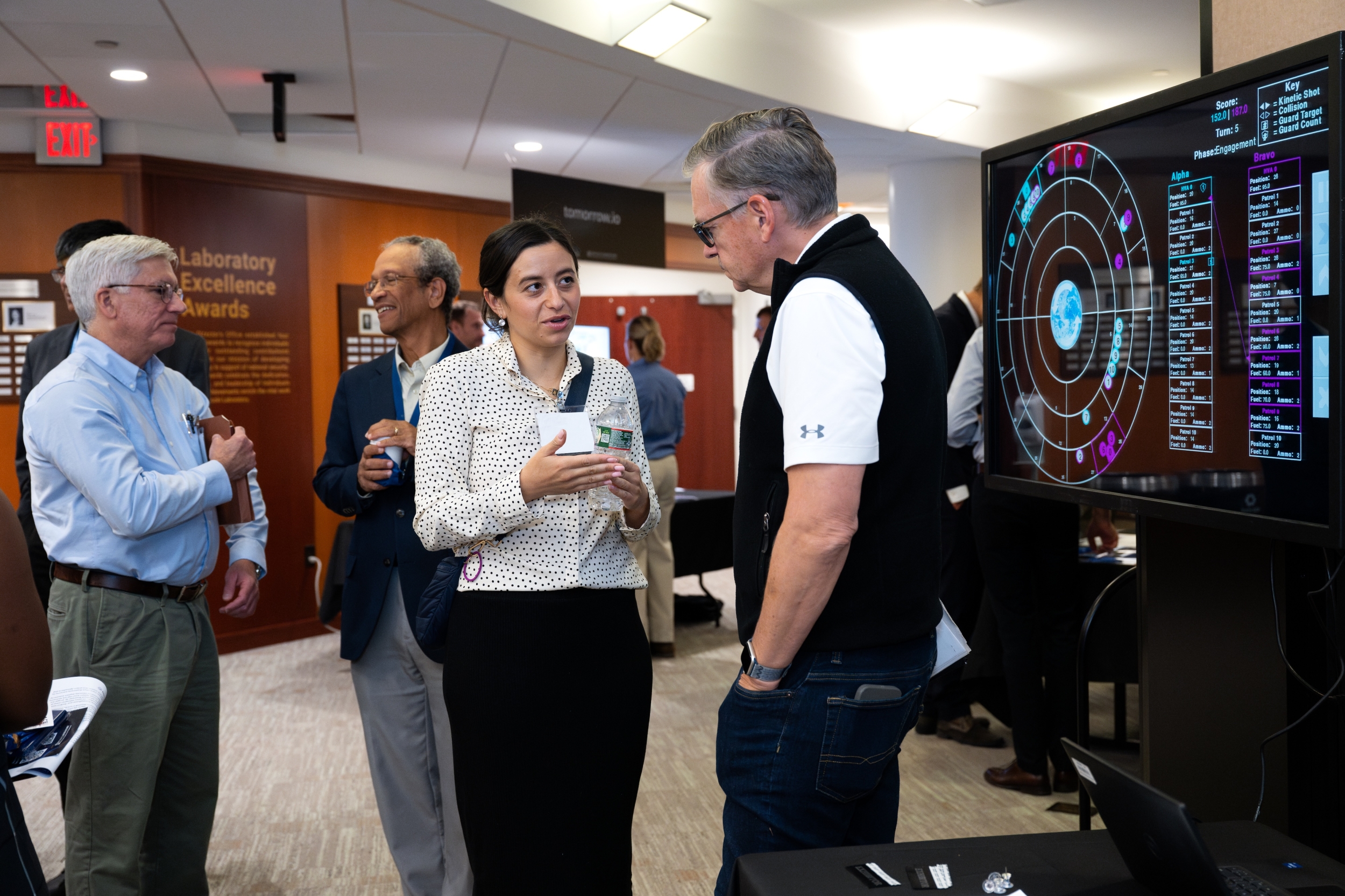 Two attendees chat in front of an exhibit shown on a screen.