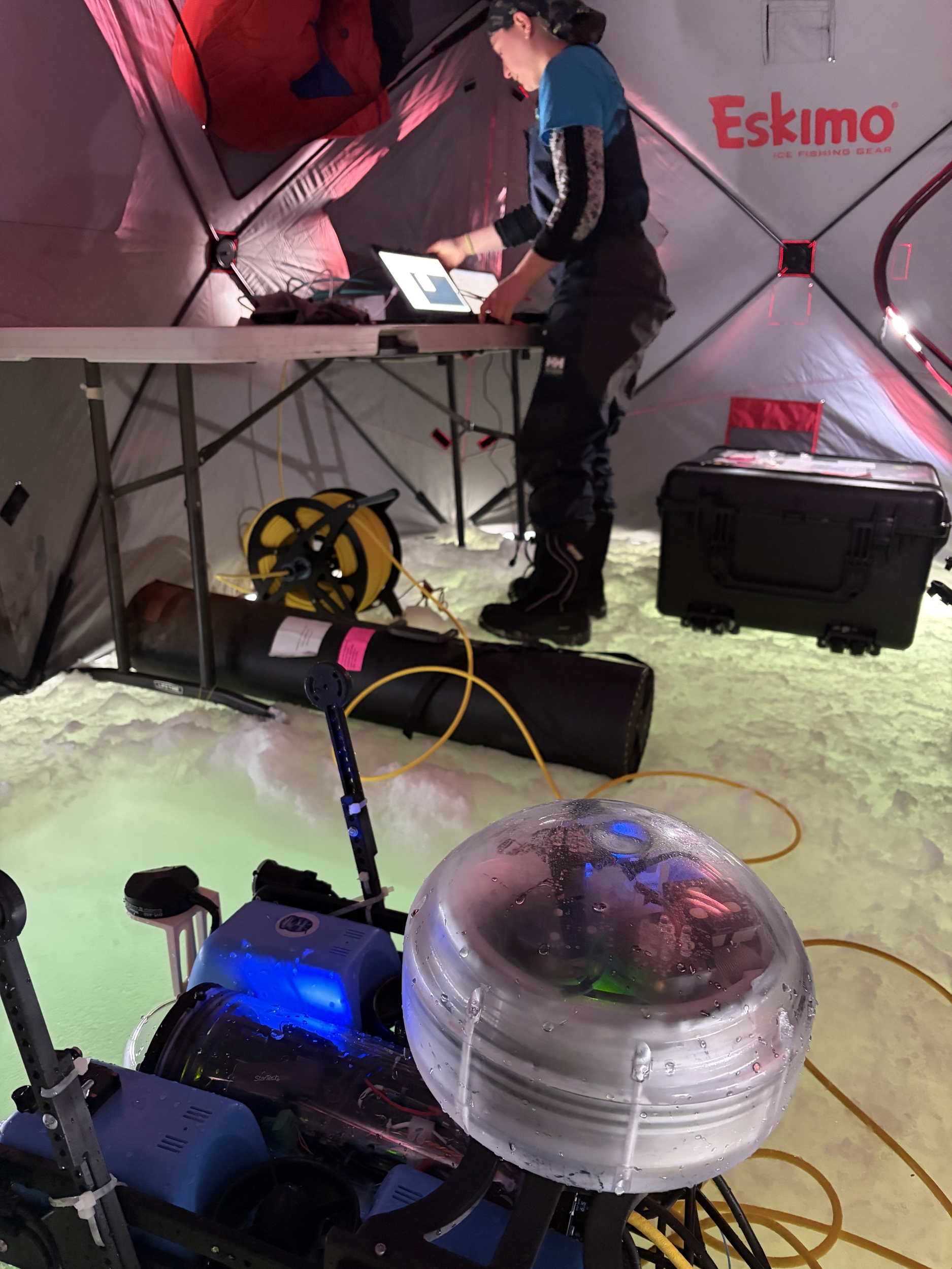 A remotely operated vehicle sits in a tent atop lagoon ice. A person in the background works on a computer.