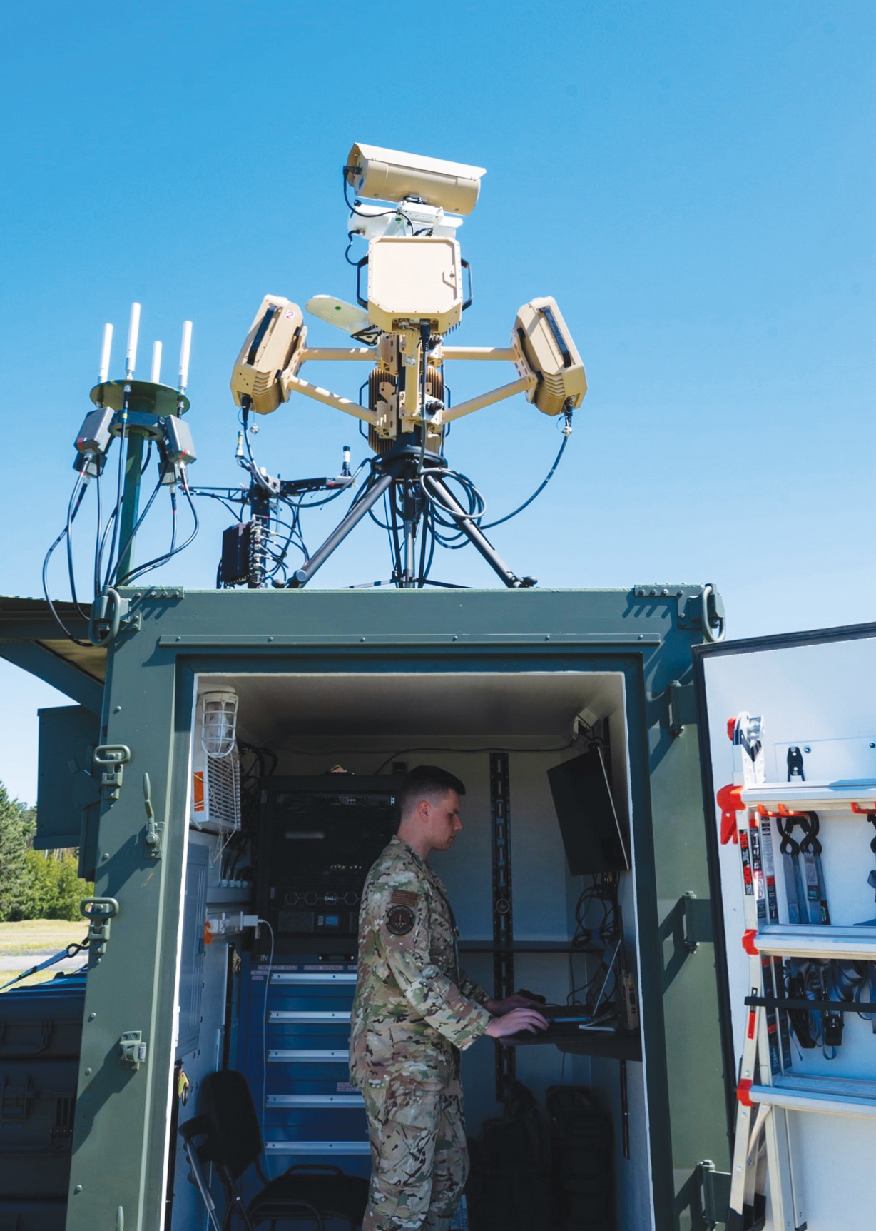 A U.S. airman stands in a small trailer operating a computer. Installed on the roof of the trailer is a system containing a camera, antennas, and electronics. 