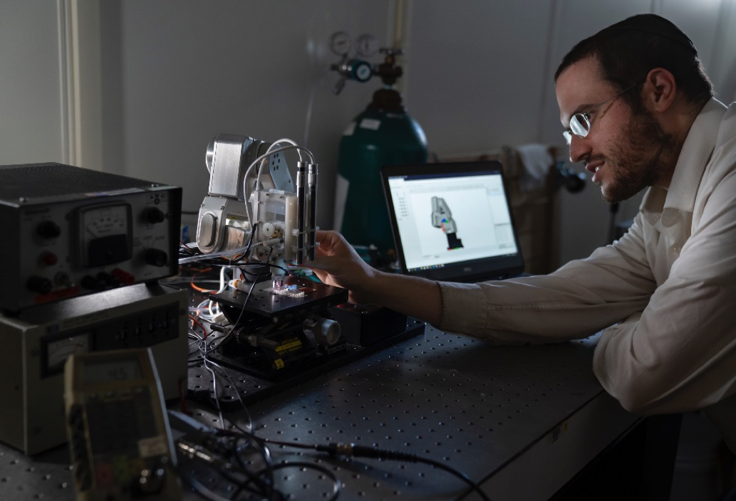 A man adjusts the microplasma sputterer setup, which looks similar to a small 3D printing arm and nozzle, depositing material onto a metal plate. 