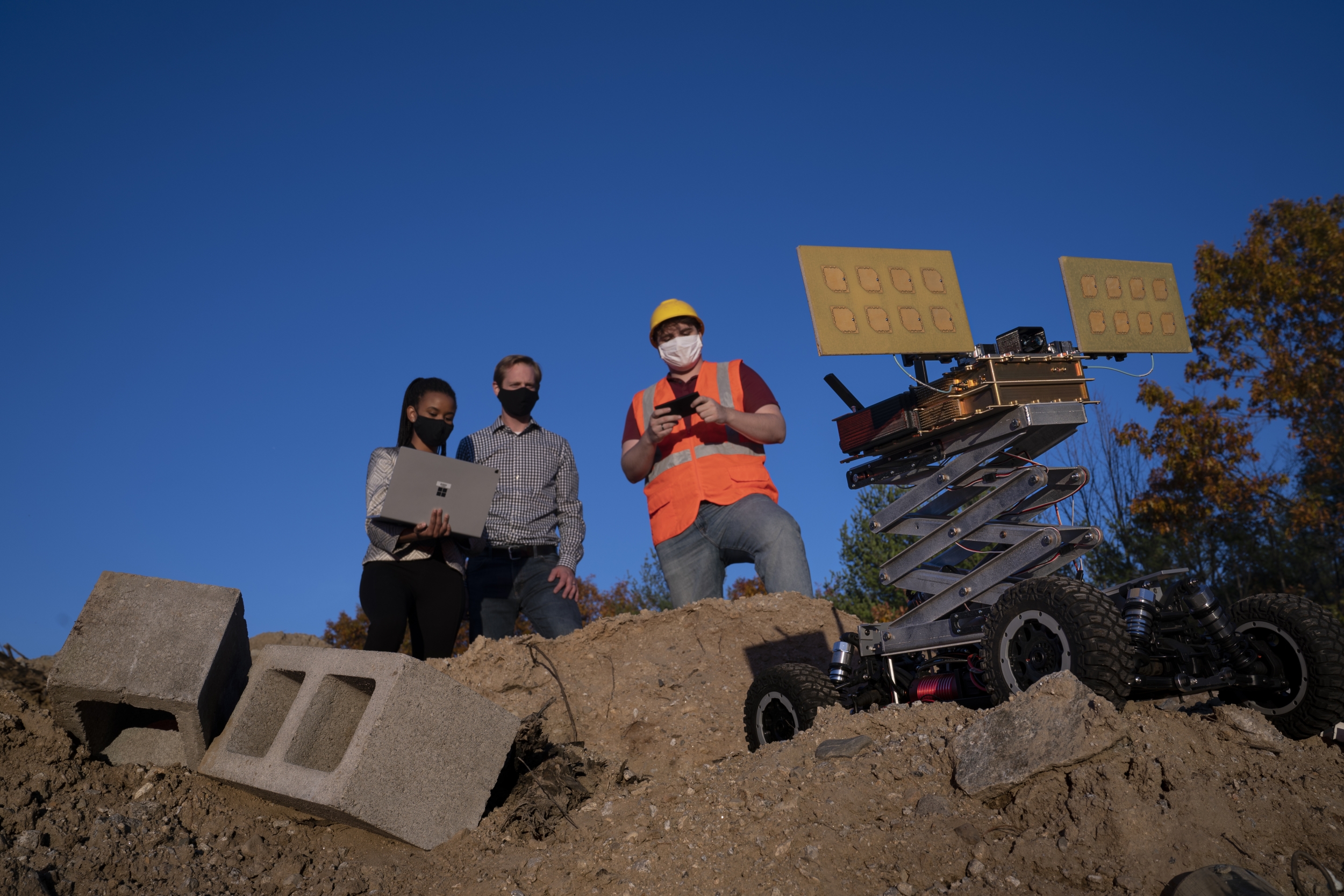 three people stand on a big dirt pile, outside, with blue sky in the background. One researcher is holding a laptop, which another looks at. The third person is holding a remote, controlling a robotic vehicle also on the dirt pile. 