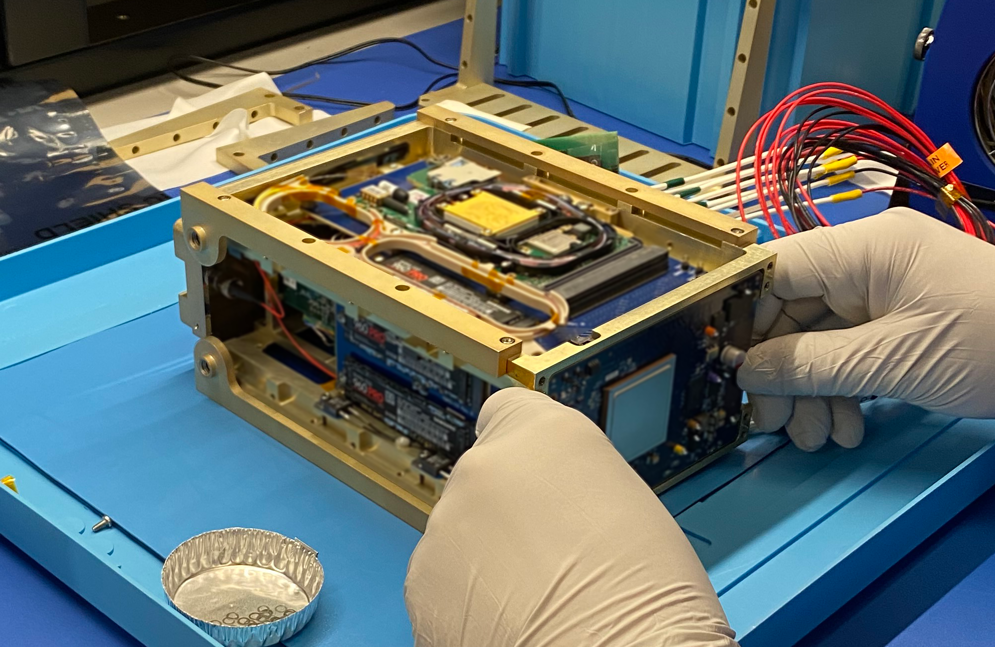 A person wearing gloves holds a tissue-box-sized communications payload, with its interior components displayed.