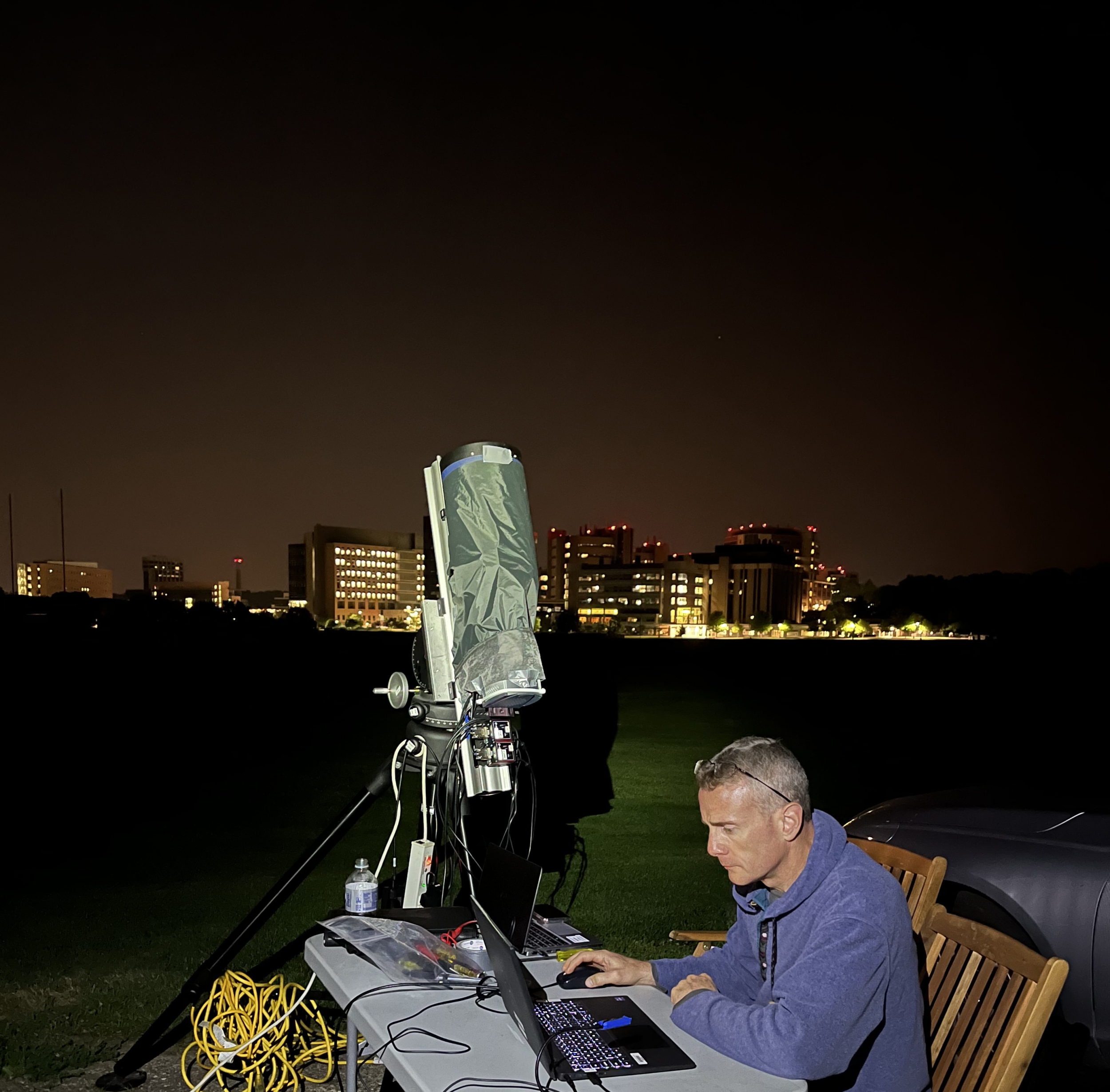 a person sits by a laptop, with a telescope beside him, pointing up to the sky. The city skyline is in the background.