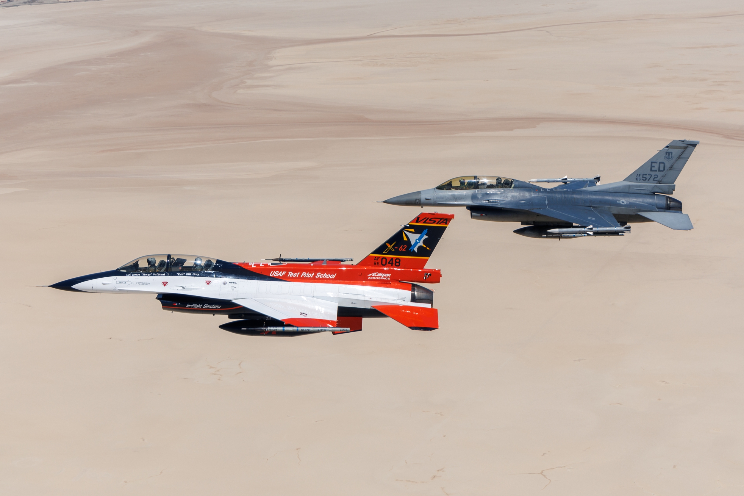 Two F-16 aircraft, one red and white and the other traditional grey, fly side by side over a desert. 