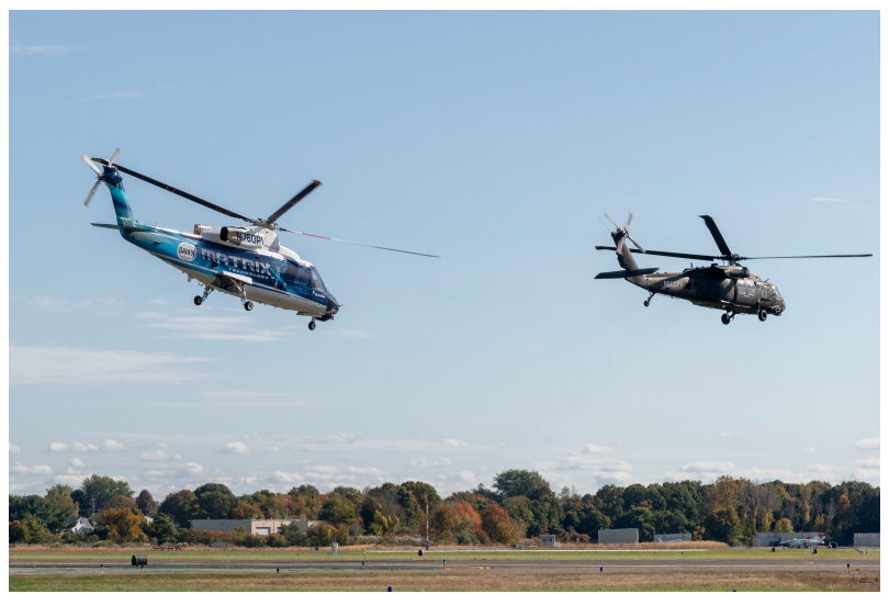 Two helicopters are shown flying at low altitude over an airfield.