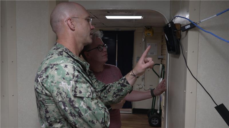 A Navy captain and a Lincoln Laboratory researcher interact with a small touchscreen installed on the wall inside of a U.S. Navy ship. 