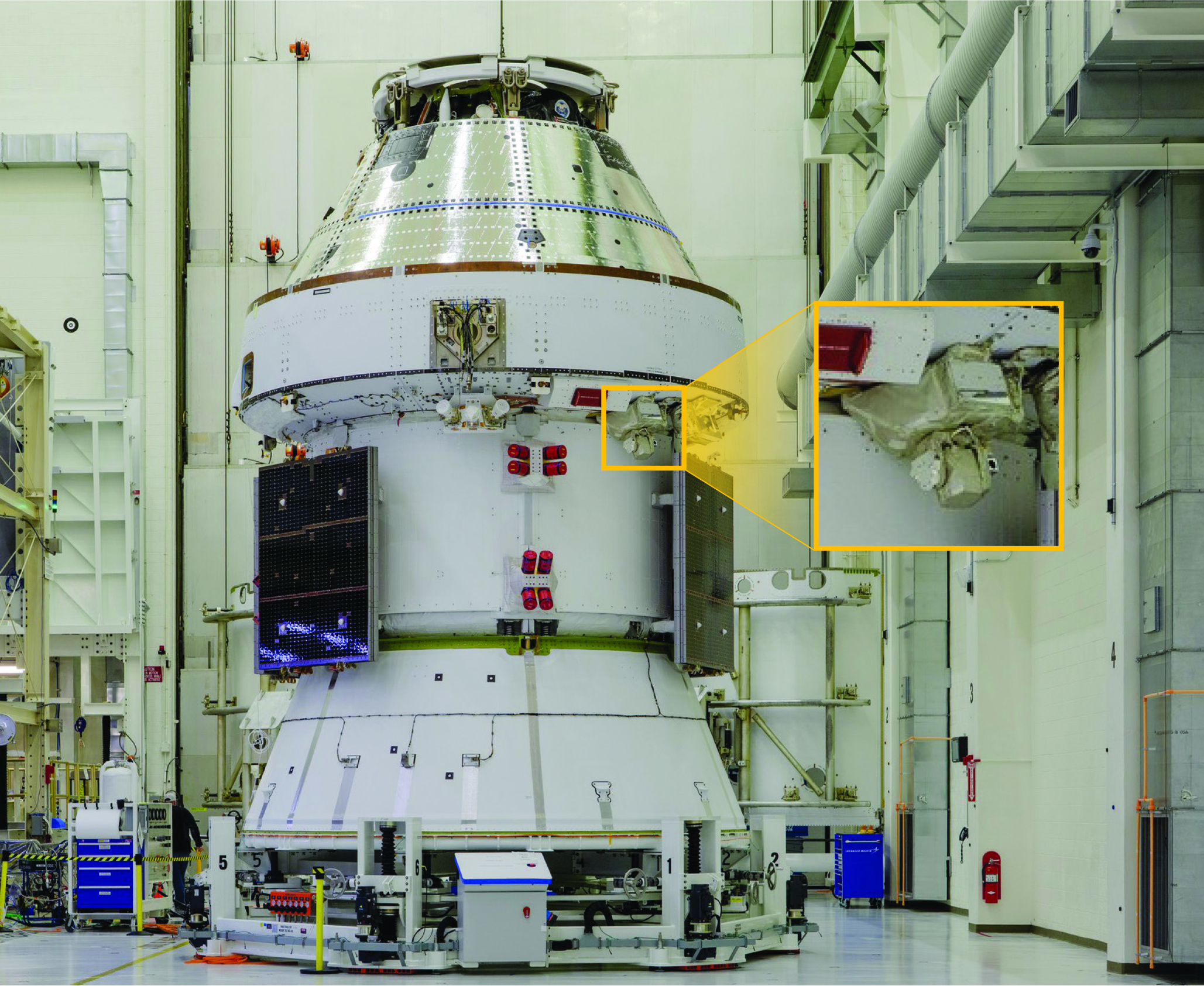 Inside of a large engineering space sits the Artemis II Orion capsule, a conical vehicle about the size of large delivery van. A photo inset shows an enlarged view of a piece of equipment on the exterior, which is the MAScOT optical terminal.