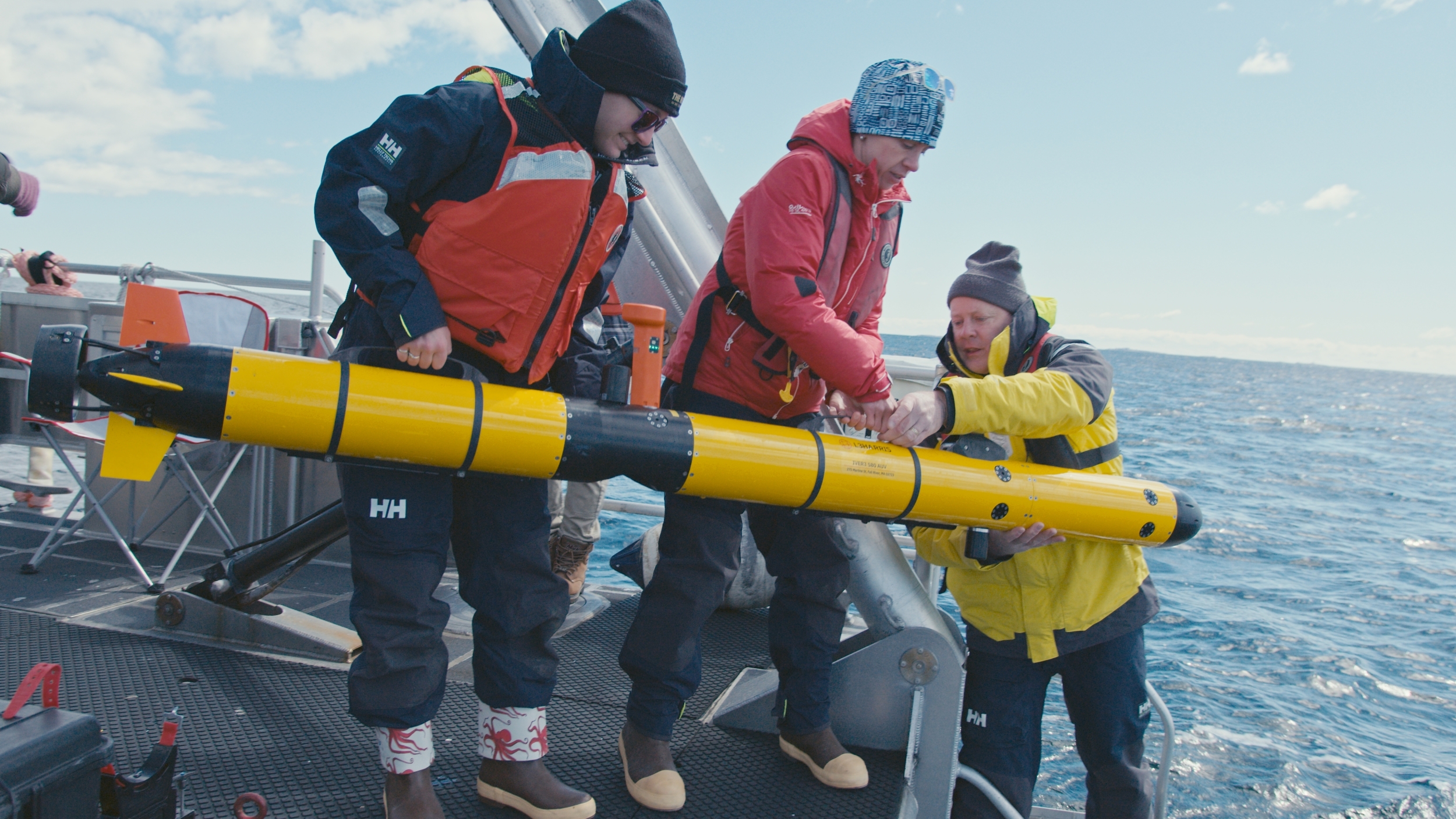 Three researchers stand on a boat deck in the ocean, holding a long skinny tube (approx 6 feet) that is an autonomous underwater vehicle. They are lowering the vehicle into the water. 
