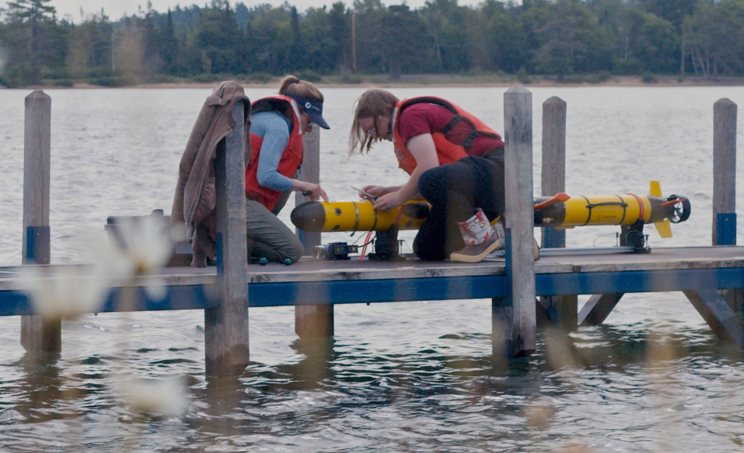 Two researchers huddle over an underwater uncrewed vehicle, making adjustments to it, on a dock over a lake. 