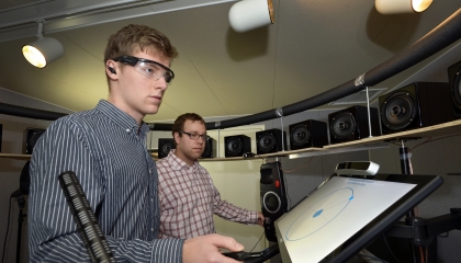 Two staff members stand in a room surrounded by speakers and use a system to select which speaker a sound is coming from.