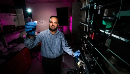 A researcher stands in a lab next to a microscope holding up a glass slide containing human tissue samples.