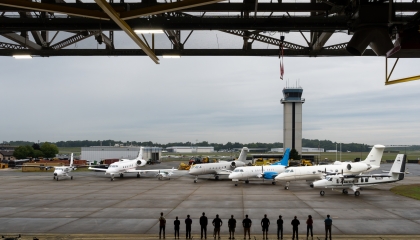 Seven aircraft line a tarmac while 12 people look out of a hangar in silhouette 
