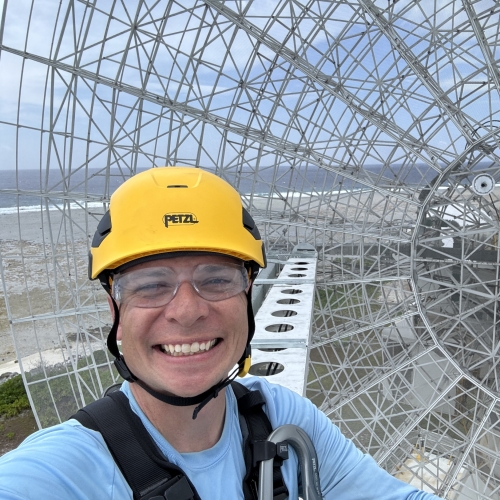 Andrew Dahir, wearing a yellow helmet, stands in front of a radar with the ocean in the backdrop.