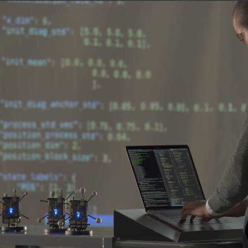 A researcher stands on a laptop. In front of him are a few small radios, and a large projector screen showing data.