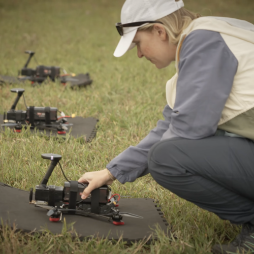 A staff member crouches beside three drones lying on the grass.