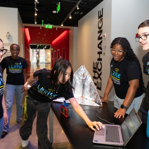 A group of LLRISE students wearing LLRISE shirts smile while preparing equipment inside of the MIT Museum.