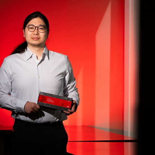 A photo of Jeffery Lim against a red background, holding a hardware platform (rectangle, about 4 by 12 inches). 