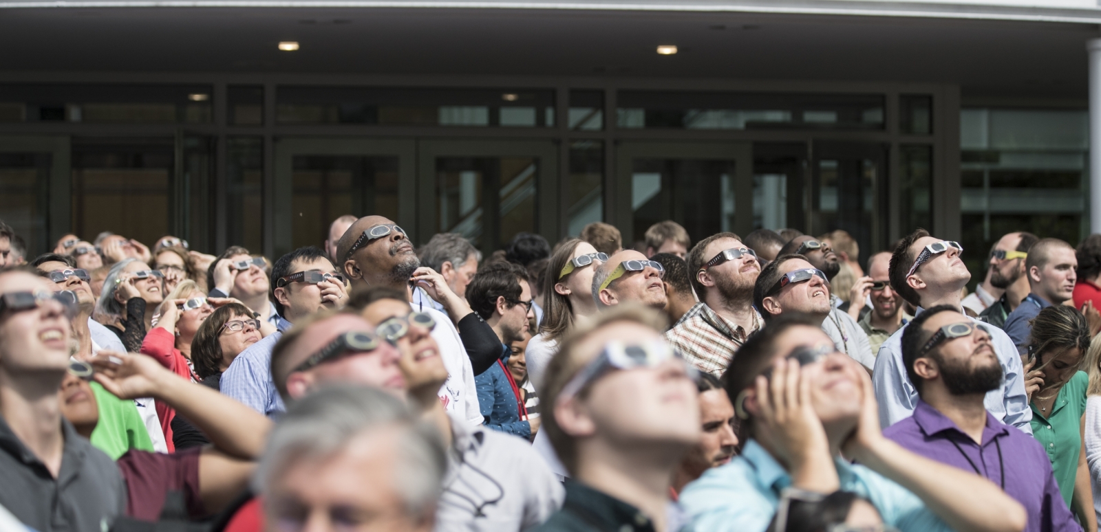 A large group of employees gather outside of the main Lincoln Laboratory entrance, each wearing special sunglasses to watch the solar eclipse.