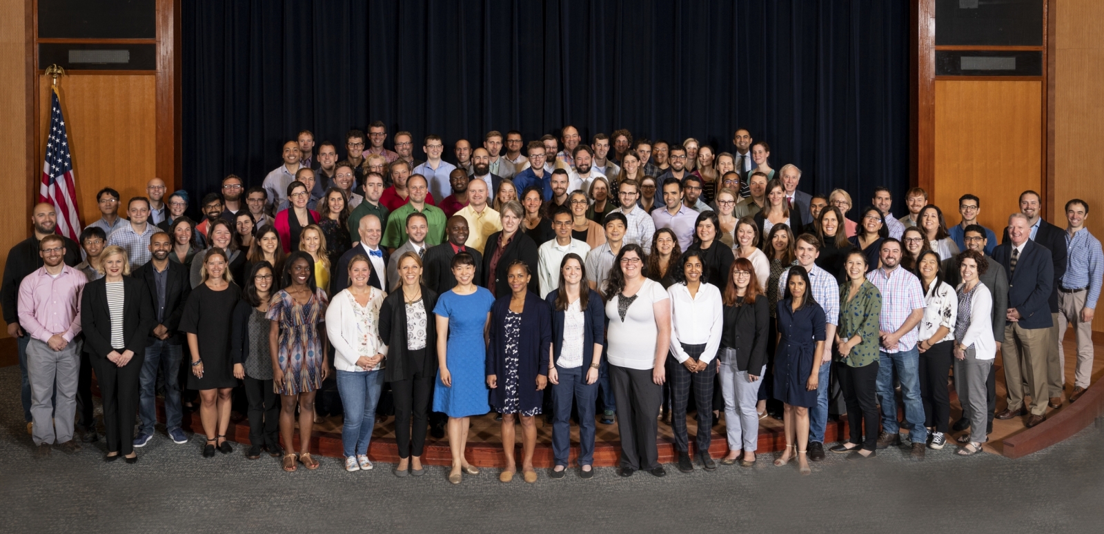 About 100 attendees of a workshop gather for a photo on an auditorium stage. 