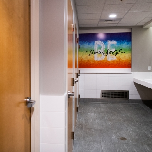 A remodeled bathroom with private closed stalls, multiple sinks, and a rainbow flag featuring the words "be yourself"